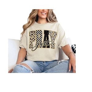 Könnte beinhalten: Beiges T-Shirt mit den Worten "PRAY just" in einer Mischung aus Leopardenmuster, schwarz-weiß karierten und schwarzen Blockbuchstaben. Das Wort "just" ist in einer Schreibschrift. Das Model trägt eine blaue Jeans.