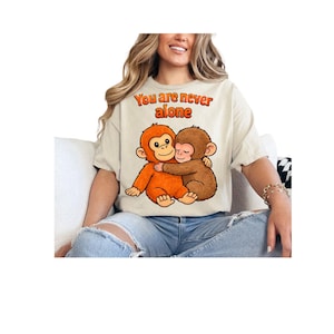 Puede incluir: Camiseta color crema con un gráfico de dos monos de dibujos animados abrazándose. El texto "You are never alone" está arqueado sobre los monos en naranja. Los monos son naranjas y marrones.