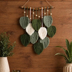 May include: A macrame wall hanging featuring leaf-shaped designs in shades of green and white. The leaves are suspended from a wooden dowel with wooden beads and a natural fiber string. The wall decor is displayed against a wooden background.