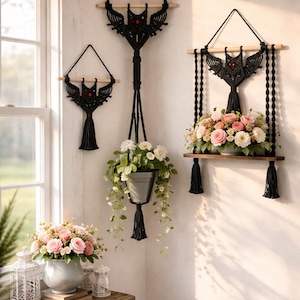May include: Black macrame plant hangers with wooden dowels and decorative tassels. The image shows three different sizes of macrame hangers, one holding a metal pot with trailing greenery and another holding a shelf with pink and white roses.