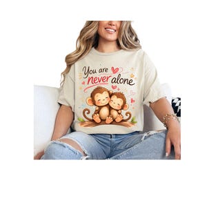 Puede incluir: Camiseta color crema con un gráfico de dos monos de dibujos animados abrazándose en una rama. El texto "You are never alone" está encima de los monos, con corazones y estrellas. La camiseta la lleva una persona.