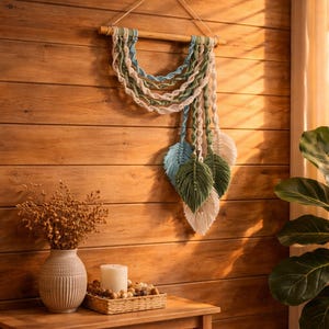 May include: A macrame wall hanging with a wooden dowel, featuring cream, blue, and green woven details. Below, three leaf-shaped accents in matching colors hang down. The wall is made of wooden planks.