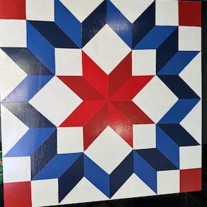 May include: A square, painted wooden wall hanging featuring a geometric star design. The design incorporates red, white, and blue hues, with a central red star and surrounding blue and white star shapes. The artwork is reminiscent of a quilt pattern.
