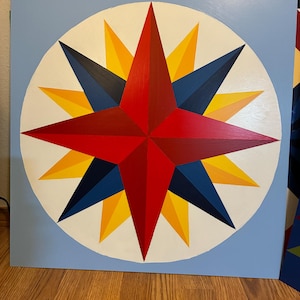 May include: A geometric star design painted on a square, light blue background. The star features red, yellow, and navy blue triangular shapes within a white circle. The artwork is displayed on a wooden surface.