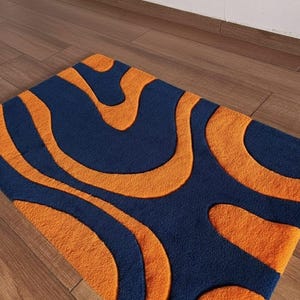 Könnte beinhalten: Ein rechteckiger Teppich mit Retro-Design. Der Teppich hat einen marineblauen Hintergrund mit orangefarbenen, wellenförmigen Mustern. Der Teppich liegt auf einem Holzboden. Die Abmessungen des Teppichs sind nicht sichtbar.