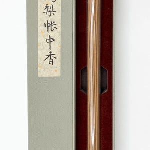 May include: Incense sticks in a clear glass tube with a gold cap, presented in a gray box with Japanese text on a label. The box has a red interior. The incense sticks are a light brown color.