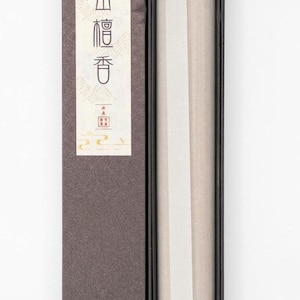 May include: A rectangular box of incense sticks, partially open, revealing the contents. The box is dark brown with a white label featuring Japanese text. A gold-colored incense holder sits atop the sticks. The incense is wrapped in white paper.