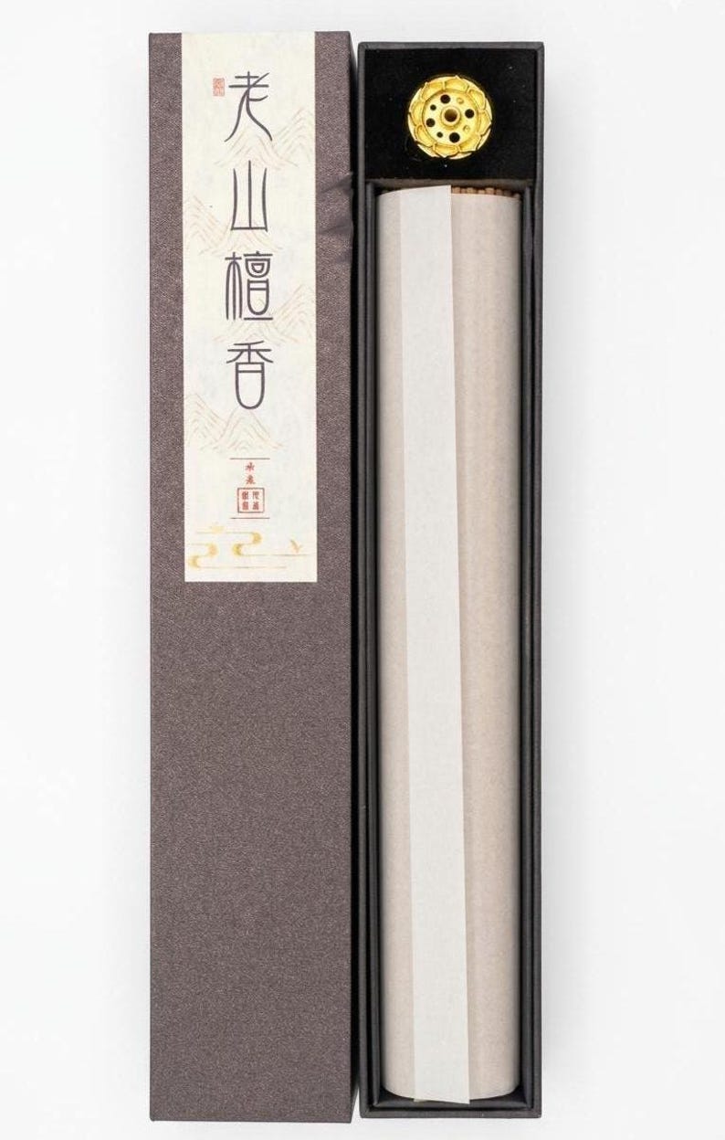 May include: A rectangular box of incense sticks, partially open, revealing the contents. The box is dark brown with a white label featuring Japanese text. A gold-colored incense holder sits atop the sticks. The incense is wrapped in white paper.