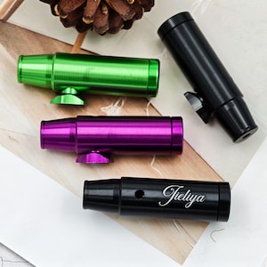 May include: A selection of metallic smoking pipes in green, purple, and black. One black pipe has the name "Tieliya" in cursive. The pipes have a cylindrical design with a flared mouthpiece and a small base. The background includes a pine cone and a white surface.