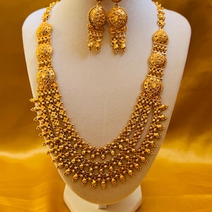 Puede incluir: Un conjunto de joyas de color dorado, que incluye un collar y pendientes a juego, exhibido en un maniquí blanco. El collar presenta múltiples hilos y detalles ornamentados. Los pendientes son redondos con diseños intrincados.