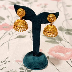 Op de afbeelding: Een paar goudkleurige jhumka-oorbellen uitgestald op een donkergroene fluwelen sieradenstandaard. De oorbellen hebben een koepelvorm met ingewikkelde filigraan en kleine bungelende kralen. De achtergrond heeft een bloemenpatroon.