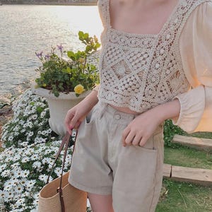 Puede incluir: Un top de ganchillo beige con cuello cuadrado y mangas cortas se combina con pantalones cortos a juego. Se sostiene un bolso tote tejido con asas de cuero marrón. El conjunto se encuentra frente a flores y agua.