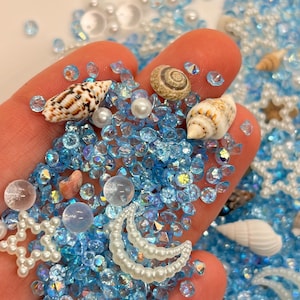 May include: A handful of blue, iridescent, and clear beads, pearls, and seashells. The beads are scattered around the hand, with a variety of shapes and sizes. The seashells are small and detailed, with a range of colors and patterns.