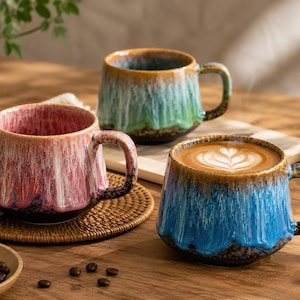 May include: Three ceramic coffee mugs with a textured, glazed finish. One mug is pink, another is blue, and the third is green. The blue mug is filled with coffee and latte art. Each mug has a handle and sits on a wooden surface.