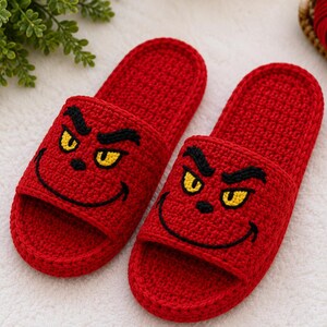 May include: A pair of red crochet slippers featuring a cartoon face design. The slippers have a slide-on style with a detailed face design in black and yellow. The slippers are made of a soft, textured material.