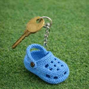 Puede incluir: Un llavero de zapato de ganchillo azul claro con un botón de bronce. El llavero está unido a una cadena plateada y a una llave dorada. El zapato en miniatura descansa sobre una superficie verde y herbosa.