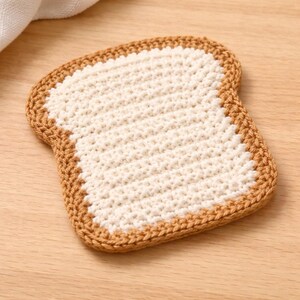 May include: A handmade, crocheted coaster shaped like a slice of bread. The coaster features a cream-colored center with a brown, textured crust. The coaster is designed to protect surfaces from hot or cold beverages.