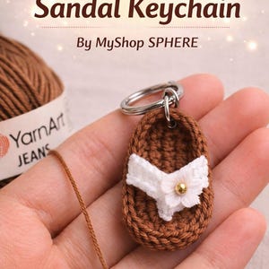 Puede incluir: Un llavero artesanal de ganchillo con forma de sandalia marrón, adornado con una flor blanca y una cuenta dorada. El llavero está sujeto a un anillo plateado. El texto "Crochet Tiny Sandal Keychain" es visible.
