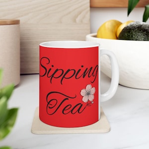 Puede incluir: Taza de cerámica blanca con fondo rojo. La taza presenta las palabras "Sipping Tea" en escritura negra, junto con una delicada ilustración de flores rosas y blancas. La taza está sobre un posavasos beige.