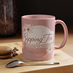 May include: A pink ceramic mug with the words "Sipping Tea minding my business" in a cursive font. The mug has a floral design and a matching pink handle. A silver spoon and a macaron are on a wooden surface.