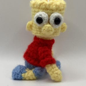 May include: A handmade crochet figure of a cartoon character with yellow skin, a red shirt, and blue shorts. The figure has large, white eyes with black pupils and is kneeling. The figure is a handmade item.