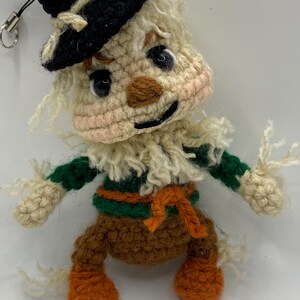 May include: A handmade crochet scarecrow doll with a black hat, cream-colored face, and orange feet. The doll has a green and brown outfit with a straw-like fringe around the neck and arms. A small metal ring is attached to the top.