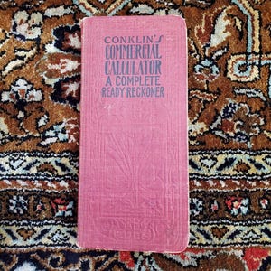Puede incluir: Un libro rosa titulado "Conklin's Commercial Calculator A Complete Ready Reckoner"