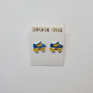 May include: A pair of blue and yellow roller skate earrings on a white card. The earrings are made of surgical steel and have yellow wheels. The card has the words "SURGICAL STEEL" printed in gold.