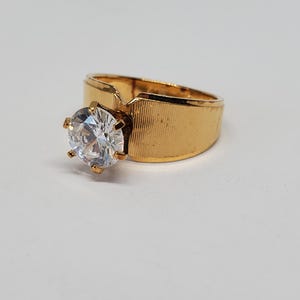 May include: A gold-coloured ring featuring a large, clear, round gemstone. The ring has a wide band with a textured surface. The gemstone is held by six prongs.