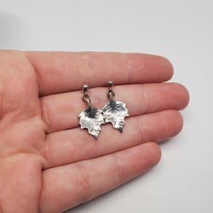 May include: A pair of silver-toned drop earrings featuring a leaf-shaped design with etched vein details. Each earring consists of a small spherical stud top connected to a dangling, textured leaf pendant with a pointed tip and serrated edges.