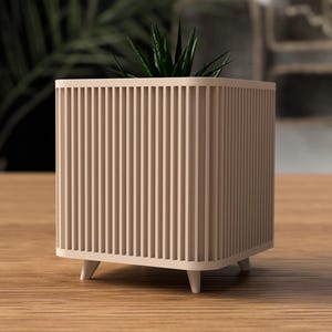 May include: A beige, square planter with vertical ribbed detailing. The planter has short, white legs and contains a green plant. The planter is on a wooden surface.