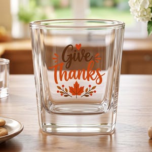 May include: A clear glass tumbler featuring the text Give Thanks in brown and orange script. The design includes a small red heart above the text and a central orange maple leaf graphic flanked by brown branches with small red berries at the base.