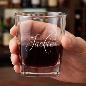 May include: A clear glass tumbler with a square base and thick walls, featuring the name Jackie etched in a white cursive script on the front surface. The glass is partially filled with a dark red liquid, showcasing its transparent design.
