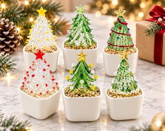 May include: Six miniature glass Christmas trees in white square pots, each filled with small pebbles. The trees feature various designs, including clear glass with red or yellow accents and green glass with colorful beads, all topped with stars.