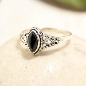 Op de afbeelding: Een zilveren ring met een centrale zwarte steen in marquise-slijpvorm. De metalen band vertoont ingewikkelde filigraan details en een kralenrand rondom de steen, wat zorgt voor een textuur langs de schacht van het sieraad.