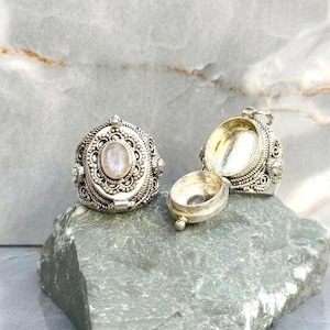 May include: A silver locket ring featuring an oval moonstone cabochon set in an ornate filigree frame. The ring is designed with a hinged compartment that opens to reveal a hollow interior, showcasing intricate metalwork and a polished finish.