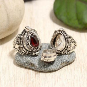 Puede incluir: Un anillo relicario de plata con un intrincado patrón de filigrana y una piedra granate de color rojo oscuro en forma de lágrima. El anillo está diseñado con un compartimento con bisagras que se abre para revelar un espacio interior hueco.