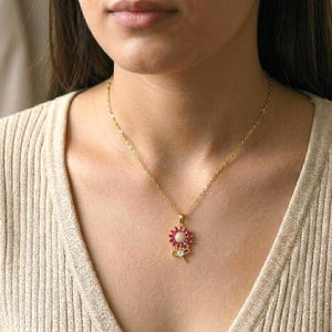 May include: A gold-colored necklace with a pendant featuring a flower design with a cluster of red gemstones and a small bow with a single white gemstone. The necklace is worn on a person.