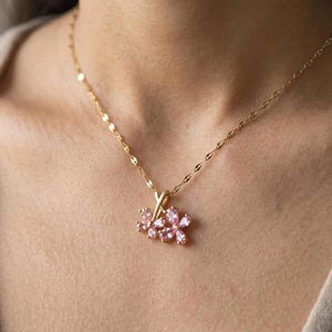 May include: A gold-colored necklace with a delicate chain and a pendant featuring three pink flower-shaped gemstones. The necklace is worn around the neck, showcasing the pendant's intricate design and color.