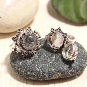 Puede incluir: Dos anillos de plata con diseño de rayos de sol. Uno presenta una piedra redonda translúcida con inclusiones negras en forma de aguja. El otro es un anillo tipo relicario abierto con un compartimento circular hueco y una tapa con bisagra.