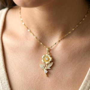May include: A gold-colored necklace featuring a flower pendant. The pendant has a central gemstone and is surrounded by petals and leaves. The chain is delicate and also gold-colored. The necklace is worn around the neck.