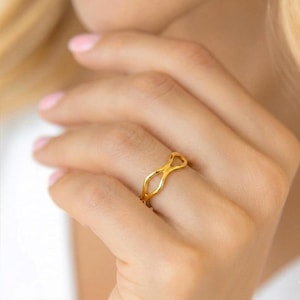 May include: A gold ring featuring an abstract infinity symbol design. The ring is worn on a finger, displaying its delicate, openwork pattern. The ring's smooth, polished surface reflects light, highlighting its warm, golden hue.