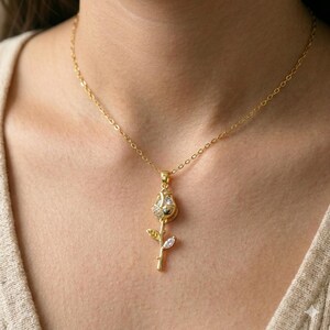 May include: A gold-colored necklace featuring a rose pendant. The rose pendant has a detailed design with clear, sparkling accents on the petals and a single, clear, teardrop-shaped accent on the stem. The necklace chain is delicate and also gold-colored.