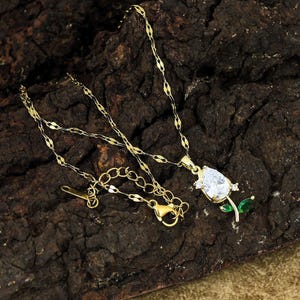 May include: Gold-colored necklace with a pendant shaped like a tulip. The pendant features a large, clear, teardrop-shaped stone and two green leaves. The necklace chain is delicate and has a lobster clasp.