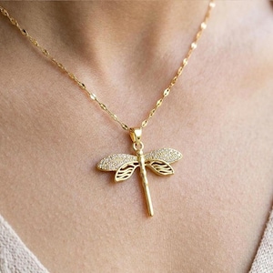 May include: A gold-colored dragonfly pendant necklace. The dragonfly has crystal-like accents on the wings. The necklace chain is a delicate, linked design. The pendant hangs against a neutral background.