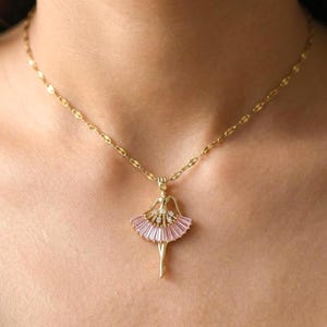 May include: A gold-colored necklace featuring a ballerina pendant. The pendant has a pink tutu, a gold-colored body, and sparkling accents. The chain is a delicate, linked design.