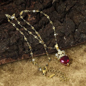 May include: A gold-colored necklace with a heart-shaped, ruby-red pendant. The pendant is topped with a small crown embellished with clear gemstones. The necklace chain has a delicate, linked design.