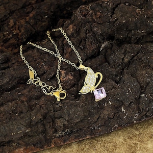 May include: A gold-colored butterfly pendant necklace. The butterfly has a wing encrusted with small, clear stones and a pink square gemstone. The necklace chain and clasp are also gold-colored.