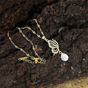 May include: A gold-colored necklace with a butterfly pendant. The butterfly wing is adorned with small, clear stones. A teardrop-shaped, clear stone hangs below the butterfly. The necklace is on a dark brown, textured surface.