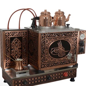 Op de afbeelding: Een koper-zwarte theemaker met twee koperen theepotten erop. De theemaker heeft sierlijke ontwerpen en een kleine koperen koffiepot. Het heeft een meter en bedieningsknoppen. Het algehele ontwerp is ingewikkeld en gedetailleerd.
